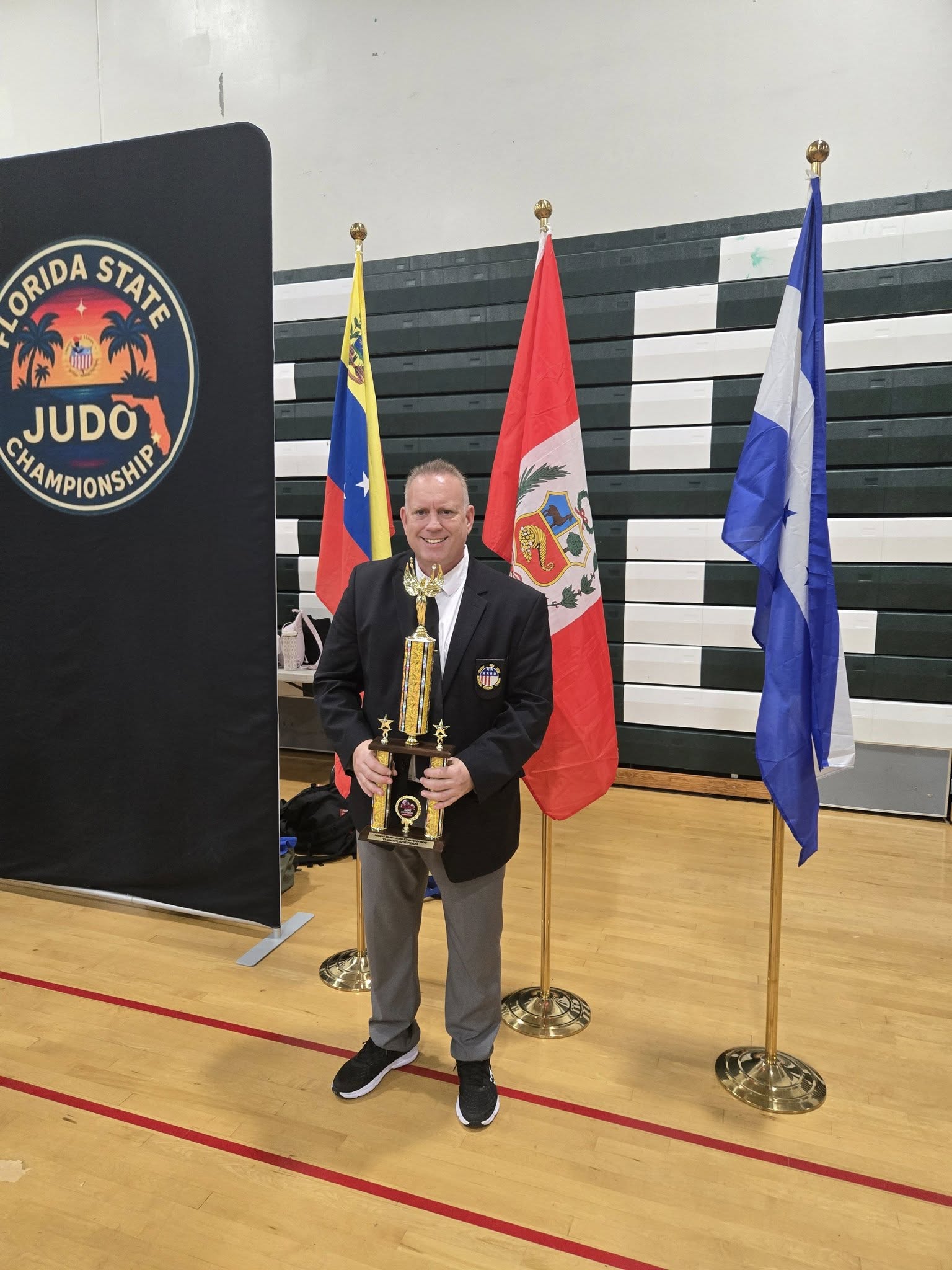 John Paccione, head coach, in a blazer with USJA pin, holding a championship trophy at the Florida State Judo Championship